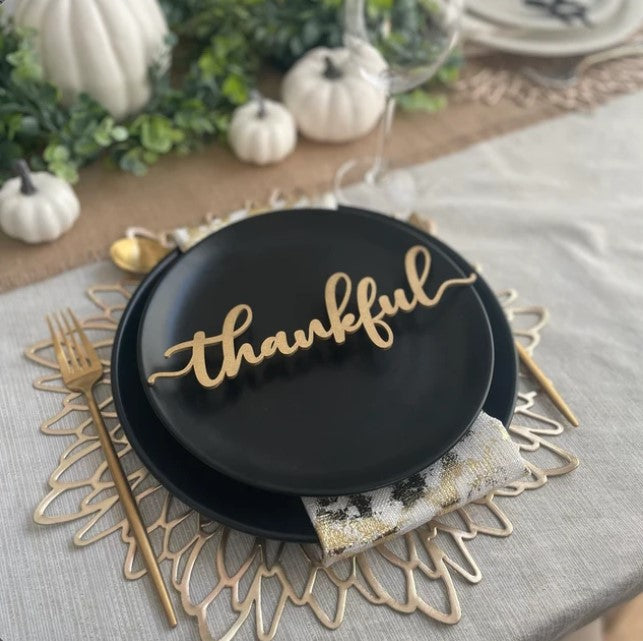 gold thankful place card on a thanksgiving table decorates with black plates gold flower charger gold silverware and a white pumpkin and greenery centerpiece