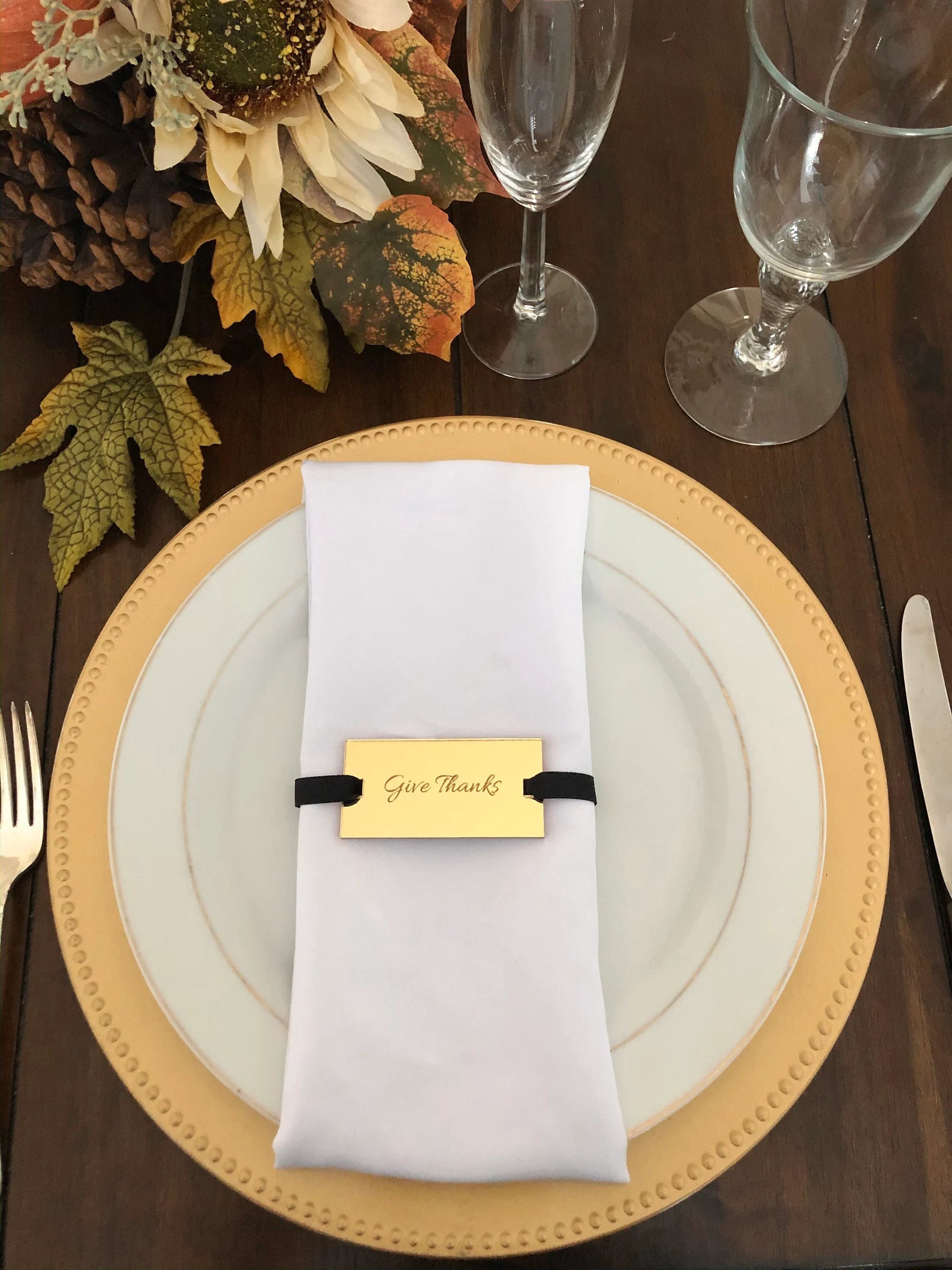Table setting with gold charger, white napkin, and gold mirror acrylic napkin tag with 'Give Thanks'  on a wooden table.