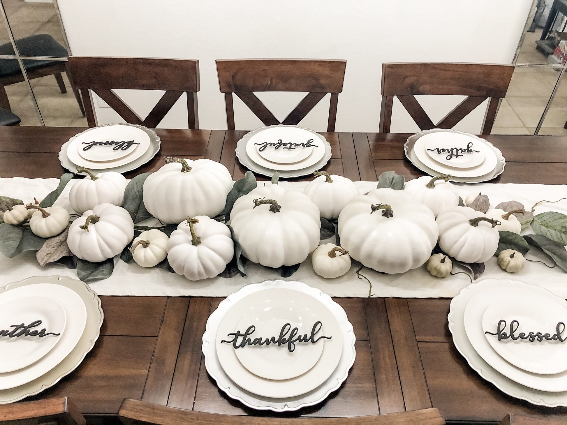 Thanksgiving table set with white plates white pumpkin centerpiece on a white table runner with green leaf garland set with thankful grateful blessed and gather place cards