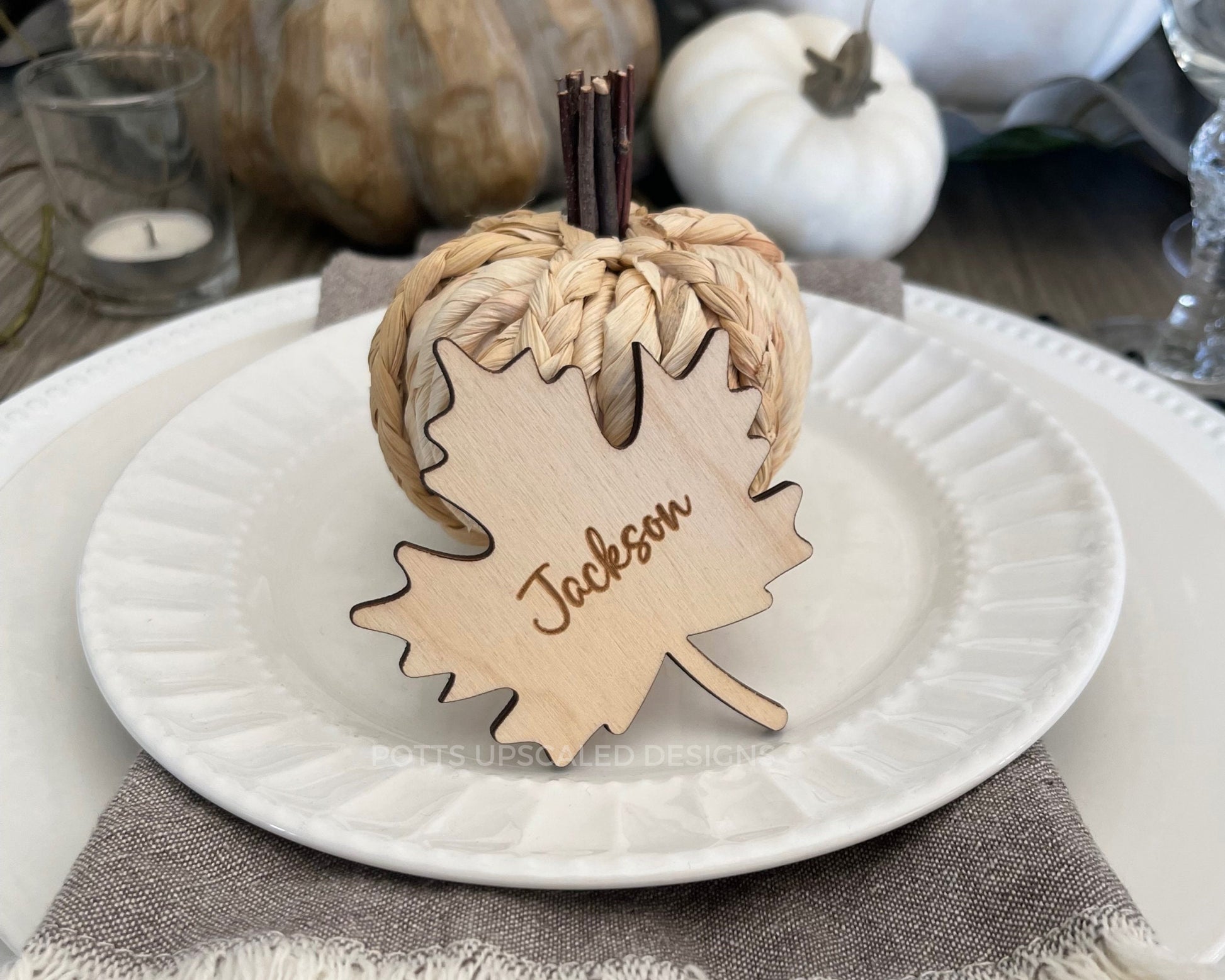 Light wood maple leaf place card engraved with guest names on a fall decorated table with white plates and wood and white pumpkins