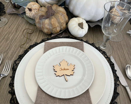 Light wood maple leaf place card engraved with guest names on a fall decorated table with white plates and wood and white pumpkins