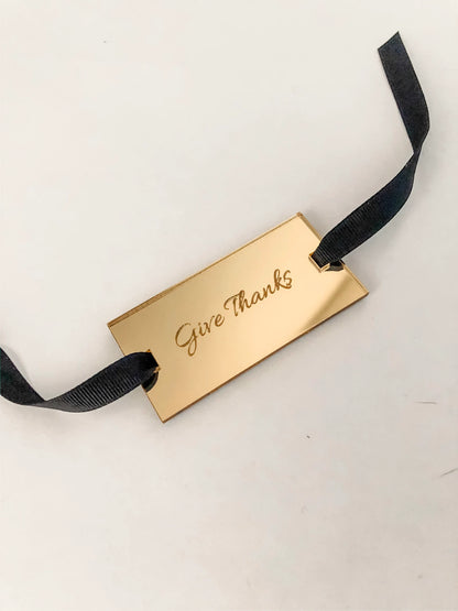 Gold mirror napkin tag with 'Give Thanks' text on a black ribbon against a white background for thanksgiving