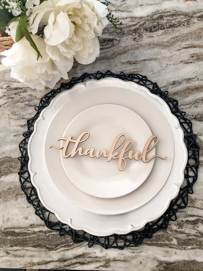 wooden thankful place card cut from light wood birch on top of a white plate, silver plate charger and black woven place mat. Thanksgiving table and white flowers