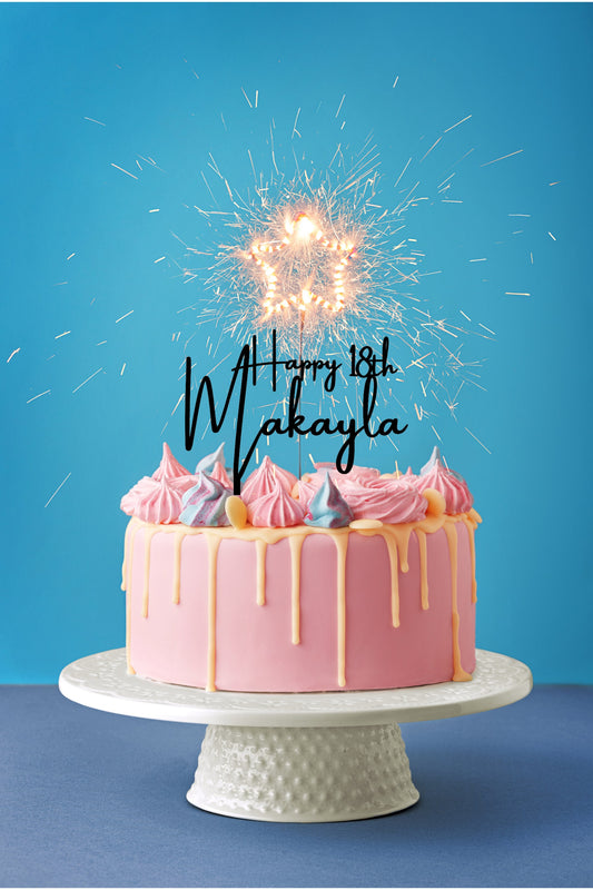 personalized happy birthday cake topper on a pink cake with a blue background.  Star sparkler candle on a white cake plate stand.  happy 18th cake topper in black