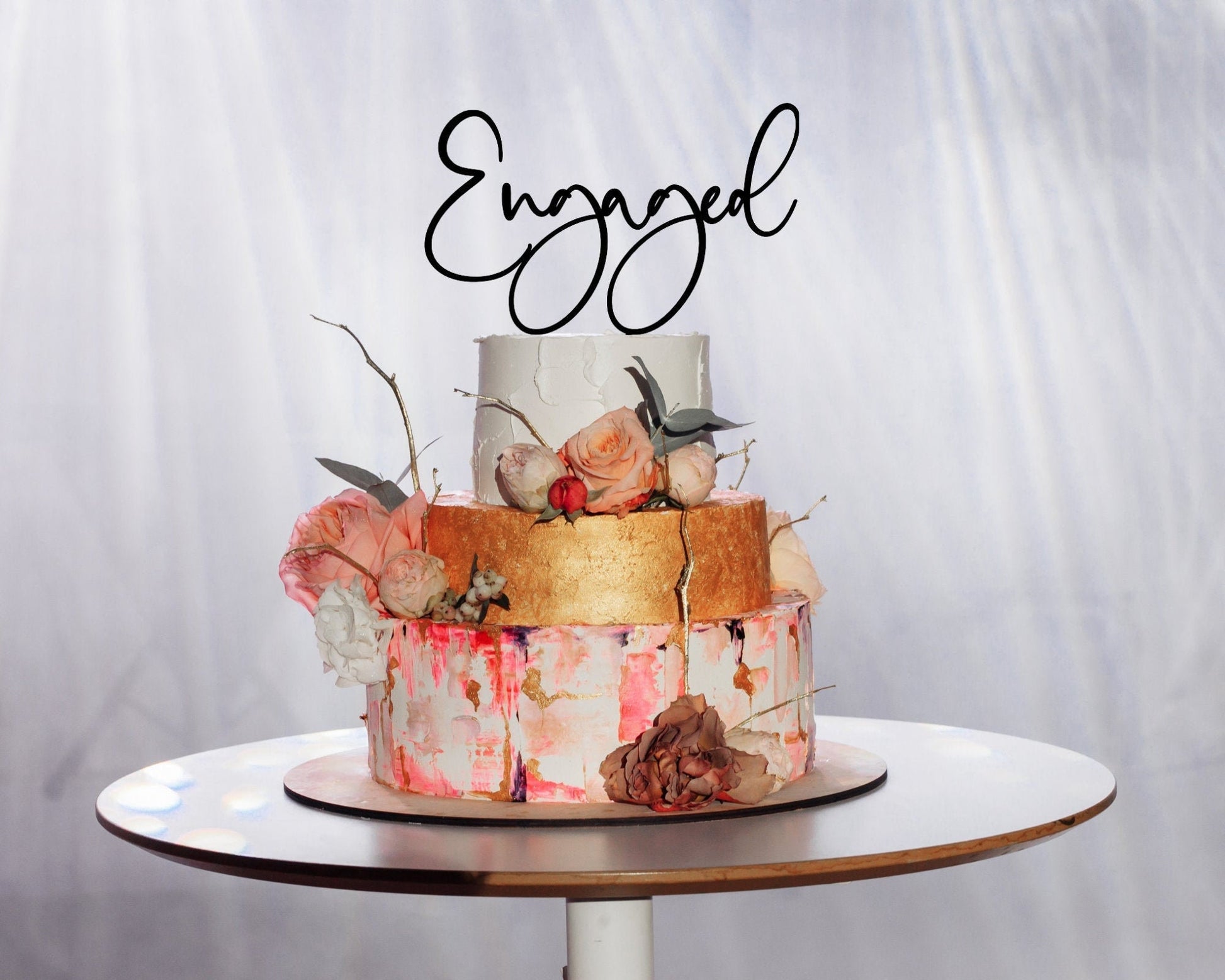 Black wood engaged cake topper in cursive font.  gold, pink and white, tiered engagement cake with roses and twigs.  Word art cake topper.