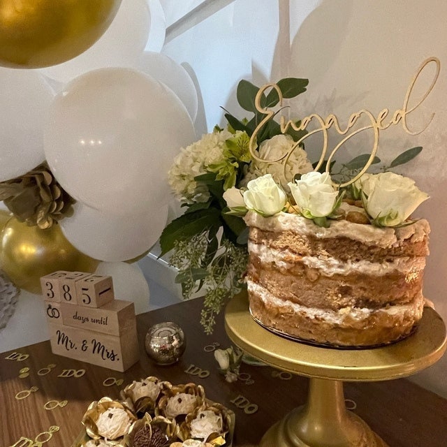 cursive font word engaged in gold wood.  Word art as a cake topper for engagement cakes.  naked layered cake with white roses and gold cake topper accent.